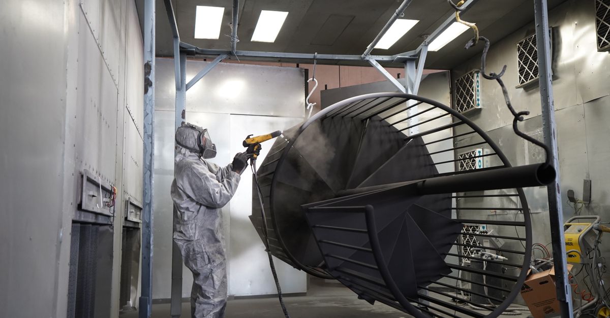 Powder-coated parts in the making: a technician in a respirator spraying black powder on a steel spiral staircase